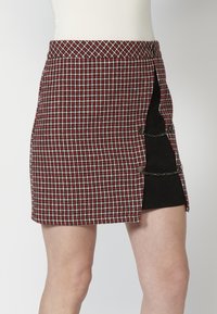 Jupe mini écossaise rouge et noire avec fentes sur les côtés, ornée de chaînes et de boutons en métal, fabriquée en tissu texturé.