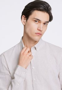 Cămașă bej deschis, cu nasturi, realizată dintr-un material texturat. Are guler, mâneci lungi și cinci nasturi pe față. Finisaj neted.