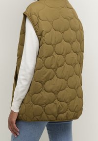 Quilted, olivgrüner ärmelloser Weste über einem weißen Langarmshirt und blauen Jeans getragen, Rücken und linke Seite sichtbar.