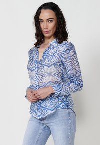 Camicia button-up blu con motivo geometrico, tessuto leggero, maniche lunghe e colletto, abbinata a jeans azzurri chiari.