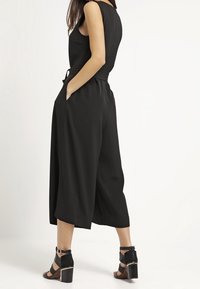 Combinaison sans manches noire avec une ceinture à la taille et un design à jambes larges, comportant des poches latérales. Associée à des sandales à talons blocs.