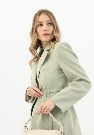 Lichtgroene getailleerde blazer met lange mouwen, een ceintuur in de taille en zijzakken. Model houdt een handtas met gevlochten handvat vast. Soepele stofstructuur.