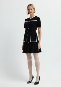 Abito nero con maniche corte, bottoni dorati, bordi bianchi e due tasche; abbinato a calze trasparenti e decorate e tacchi alti neri.