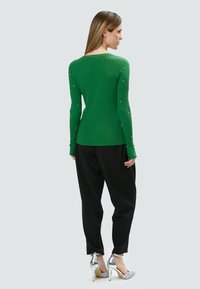 Maglione lungo a maniche lunghe verde a coste con piccoli dettagli decorativi sulle maniche, abbinato a pantaloni nero larghi e tacchi a stiletto argentati.