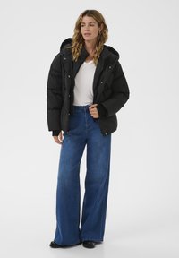 Schwarze Daunenjacke mit Kapuze, ausgestattet mit einem Reißverschluss und Knopfdetails, getragen über einem weißen Shirt und kombiniert mit weit geschnittenen blauen Jeans.