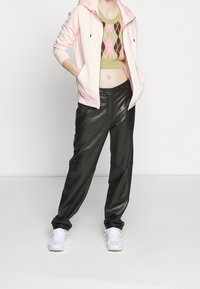 Ljusrosa zip-up hoodie, kort i modellen, över en grön argyle-mönstrad tröja. Svarta läderbyxor och vita sneakers fulländar outfiten.