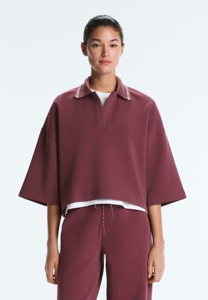 Žena oblečená v voľnom bordovom pulóvri s krátkymi rukávmi a golierom a zladených nohaviciach, stojaca pred jednoduchým bielym pozadím.