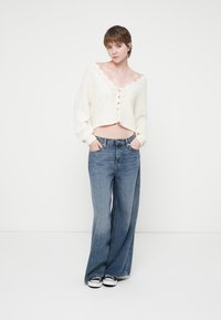 Weißer, gekürzter Pullover mit Knopfverschluss und Spitzenbesatz, kombiniert mit blauen, weit geschnittenen Jeans und schwarz-weißen Sneakers.