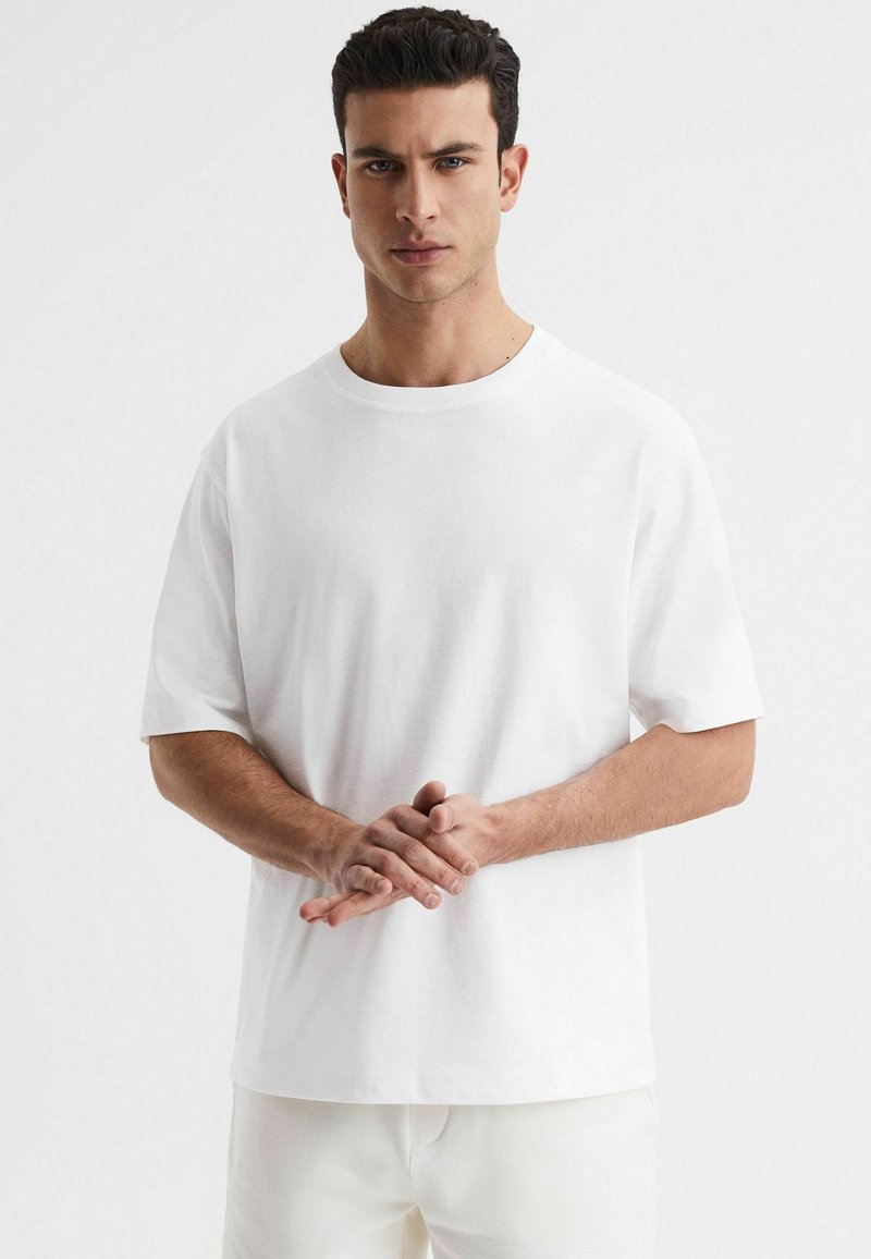T-shirt bianco in cotone con collo rotondo, maniche corte, vestibilità rilassata e texture liscia. Senza stampe visibili o dettagli.