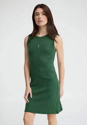 Kobieta o długich brązowych włosach, ubrana w zieloną, bezrękawnikową, prążkowaną sukienkę, stojąca na tle jasnego, jednolitego tła.