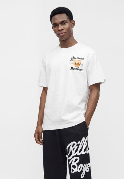 Νέος άντρας που φοράει λευκό t-shirt Billionaire Boys Club και μαύρα σορτς με μεγάλο λευκό κείμενο, στέκεται μπροστά σε ένα απλό φόντο.