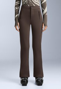 Kobieta ubrana w dopasowane brązowe spodnie z przednimi kieszeniami na zamek i czarne buty narciarskie na jednolitym jasnym tle.