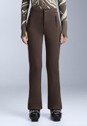 Nő szűk barna nadrágot visel elülső cipzáras zsebekkel és fekete síbakancsokkal, egyszínű világos háttér előtt.