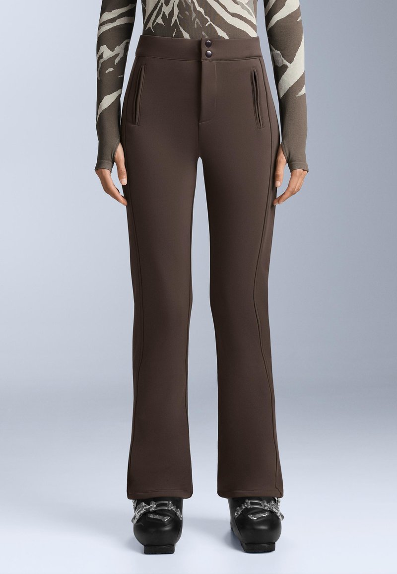 Kobieta ubrana w dopasowane brązowe spodnie z przednimi kieszeniami na zamek i czarne buty narciarskie na jednolitym jasnym tle.