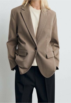 Kobieta ubrana w brązową kratkę marynarkę na białej koszuli, w połączeniu z luźnymi czarnymi spodniami, stoi z rękami w kieszeniach.