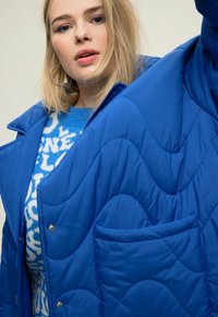 Giacca trapuntata blu con motivo a onde, dotata di chiusura a bottoni e tasca sul petto. Indossata sopra un maglione blu e bianco a motivi.
