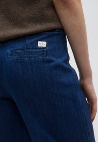 Σκούρο μπλε τζιν με πίσω τσέπη και άσπρη ετικέτα με ανάγλυφο το λογότυπο "Pepe Jeans." Το ύφασμα έχει ομαλή υφή και ελαφριά γυαλάδα.