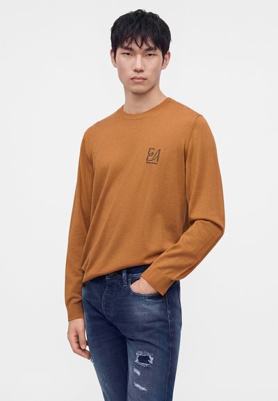 Jeune homme aux cheveux noirs portant un pull marron à manches longues avec un logo et un jean bleu usé, debout avec une main dans la poche.