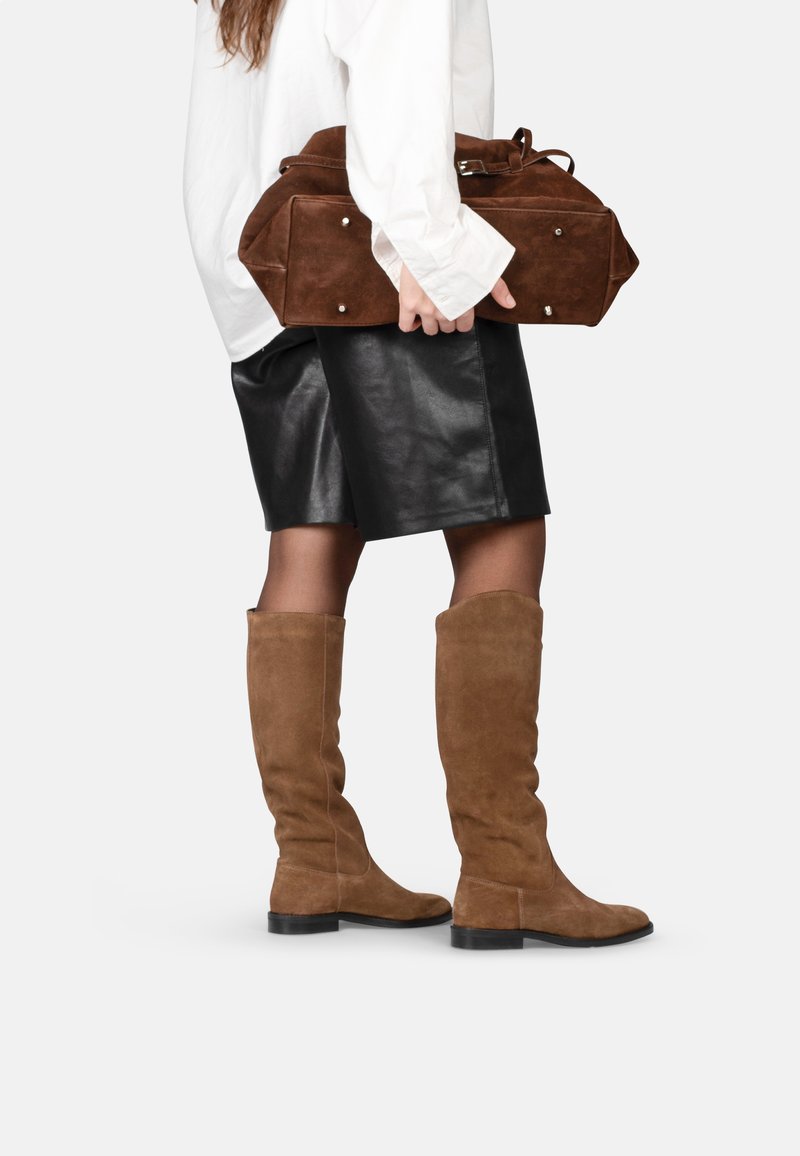 Stivali oltre il ginocchio in camoscio marrone con suola piatta, indossati con una gonna in pelle nera e una borsa in camoscio marrone, tenuta in una mano.