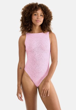 Vrouw met krullend haar, gekleed in een mouwloos lichtroze textuurrijk bodypak, staand met de ene hand ontspannen langs haar zijde en de andere bij haar dij.