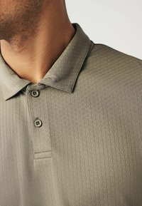 Helles olivgrünes Polohemd mit strukturiertem Muster, das einen Kragen und eine Knopfleiste mit zwei Knöpfen aufweist.