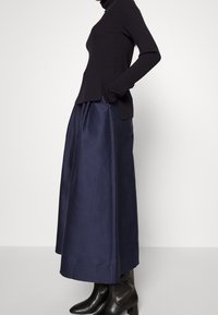 Haut à col roulé noir côtelé à manches longues, associé à une jupe fluide bleu marine, en tissu texturé, et des bottines noires à talons montant à la cheville.