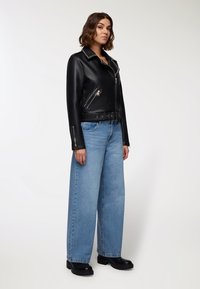 Giacca da biker in pelle nera con borchie argentate, tasche con zip e design accorciato. Abbinata a jeans azzurri a gamba larga e stivali neri.