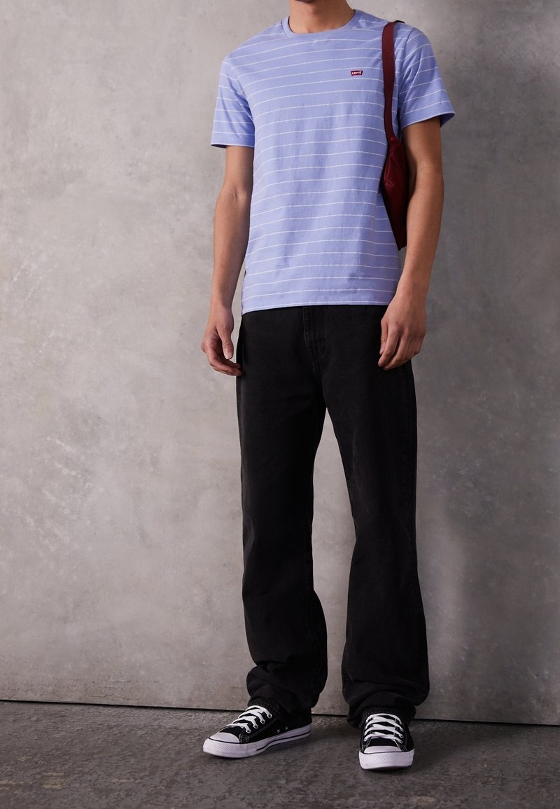 Personne portant un t-shirt à rayures bleu clair, un jean noir, des baskets noires avec des lacets blancs et un sac à bandoulière rouge devant un mur gris.