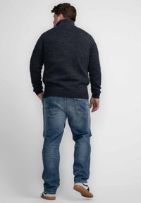 Donkergrijze, gestructureerde gebreide trui met een hoge kraag, gecombineerd met blauwe spijkerbroek en bruine schoenen, gezien vanaf de achterkant.