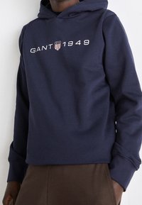 Persona vestida con una sudadera azul marino con "GANT 1949" y un logo de escudo en el pecho, combinada con pantalones marrones sobre un fondo liso.