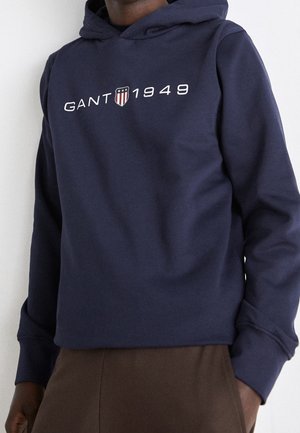 Persoon in een marineblauwe hoodie met "GANT 1949" en een schildlogo op de borst, gecombineerd met bruine broek tegen een effen achtergrond.