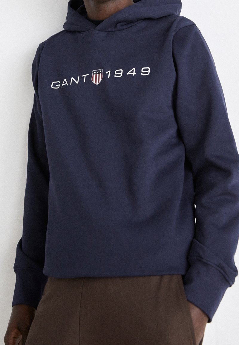 Persona vestida con una sudadera azul marino con "GANT 1949" y un logo de escudo en el pecho, combinada con pantalones marrones sobre un fondo liso.
