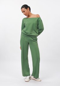 Sweatshirt verde con spalle scoperte abbinato a pantaloni larghi coordinati, entrambi in un tessuto liscio. Sneakers con strisce nere completano l'outfit.