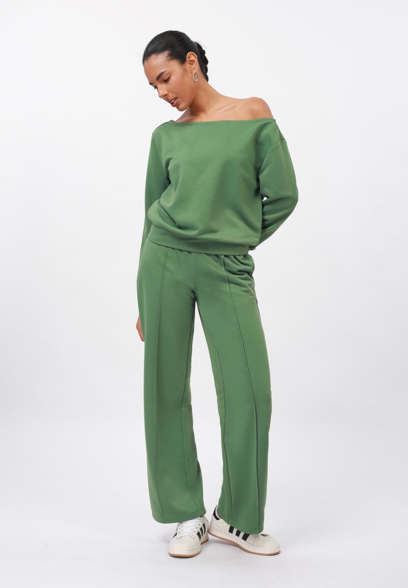 Sweatshirt verde con spalle scoperte abbinato a pantaloni larghi coordinati, entrambi in un tessuto liscio. Sneakers con strisce nere completano l'outfit.