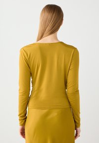 Langslieve mosterdgele top met een ronde hals en een soepele textuur. Subtiele naaideetails langs de schouders en de rug.