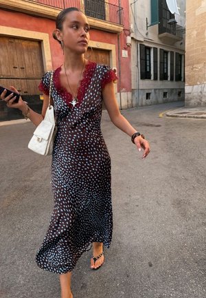 Жена, която върви по улицата, облечена в черна рокля с сини точки, червен дантелен деколте, държи телефон и бяла чанта.