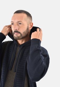 Maglione con cappuccio di colore blu scuro, lavorato a maglia e con una texture a coste, arricchito da sezioni di un verde oliva a contrasto. Presenta un cordino regolabile e una vestibilità comoda.