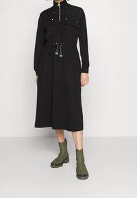 Black long-sleeve dress with a high collar, two chest pockets, drawstring waist, and a straight skirt. Worn with green ankle boots.