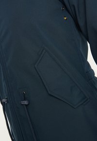 Veste bleu marine à texture lisse, dotée d'une poche en forme de losange, d'une taille à cordon de serrage et d'accents à clous près de l'épaule.