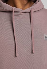 Light mauve hoodie made of textured fabric with a front pocket, drawstring hood, and a small logo patch on the chest.