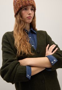 Grüne Wollstrickjacke mit gerippten Bündchen, zwei schwarzen Knöpfen, getragen über einem blauen Denimhemd mit Druckknöpfen. Das Modell hat welliges Haar und trägt eine Strickmütze.