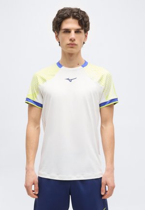 Νέος άνδρας που φοράει λευκό αθλητικό πουκάμισο με κίτρινες ρίγες στα μανίκια και μπλε περιθώριο, σε συνδυασμό με σκούρο μπλε σορτς, κοιτάζοντας προς τα εμπρός μπροστά σε απλό φόντο.