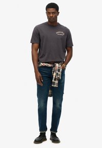 Grå bomulds t-shirt med "Superdry Los Angeles" tryk, sammen med blå jeans og en ternet flannel skjorte bundet om taljen. Sorte sko.