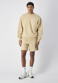 Sudadera de cuello redondo beige y pantalones cortos a juego, hechos de una tela suave, con un corte relajado y puños acanalados. Combinado con zapatillas deportivas blancas.