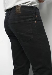 Person in schwarzen Jeans mit einem Lederaufnäher nahe dem Bund und kupfernen Nieten an den Taschenkanten.