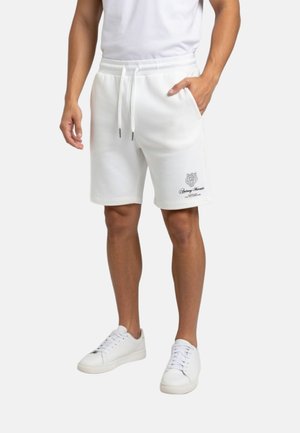 Hombre con pantalones cortos blancos con cordón y bolsillos laterales, y zapatillas blancas, de pie frente a un fondo liso.