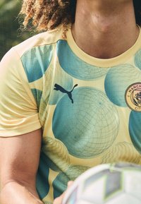 Krátký rukáv žlutého dresu s modrými kulatými vzory, s černým logem Puma a znakem fotbalového klubu Manchester City na hrudi.