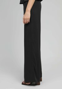 Femme portant un pantalon large noir et des sandales à talons ouverts marron, se tenant contre un fond gris clair uni.