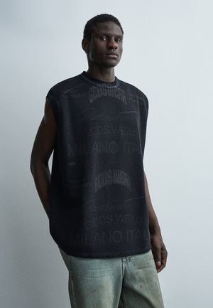 Νέος άνδρας που φοράει μαύρο αμάνικο μπλουζάκι με σχέδιο και ανοιχτό μπλε τζιν, στέκεται μπροστά σε ένα απλό ανοιχτό γκρι φόντο.