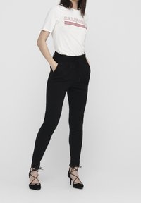 Witte T-shirt met rode tekst "CALIFORNIA", gecombineerd met zwarte broek met elastische tailleband, voorzien van zijzakken en zwarte hoge hakken.